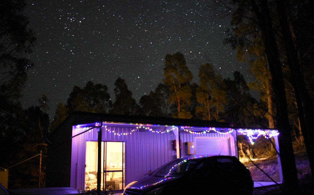 An image of the shed at night, with warm lighting coming from inside as the exterior is draped in bright Christmas lights. overhead is a clear starry night.