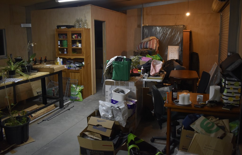 Photo shows the interior of the shed, piled with boxes, bags and the occasional pot plant.